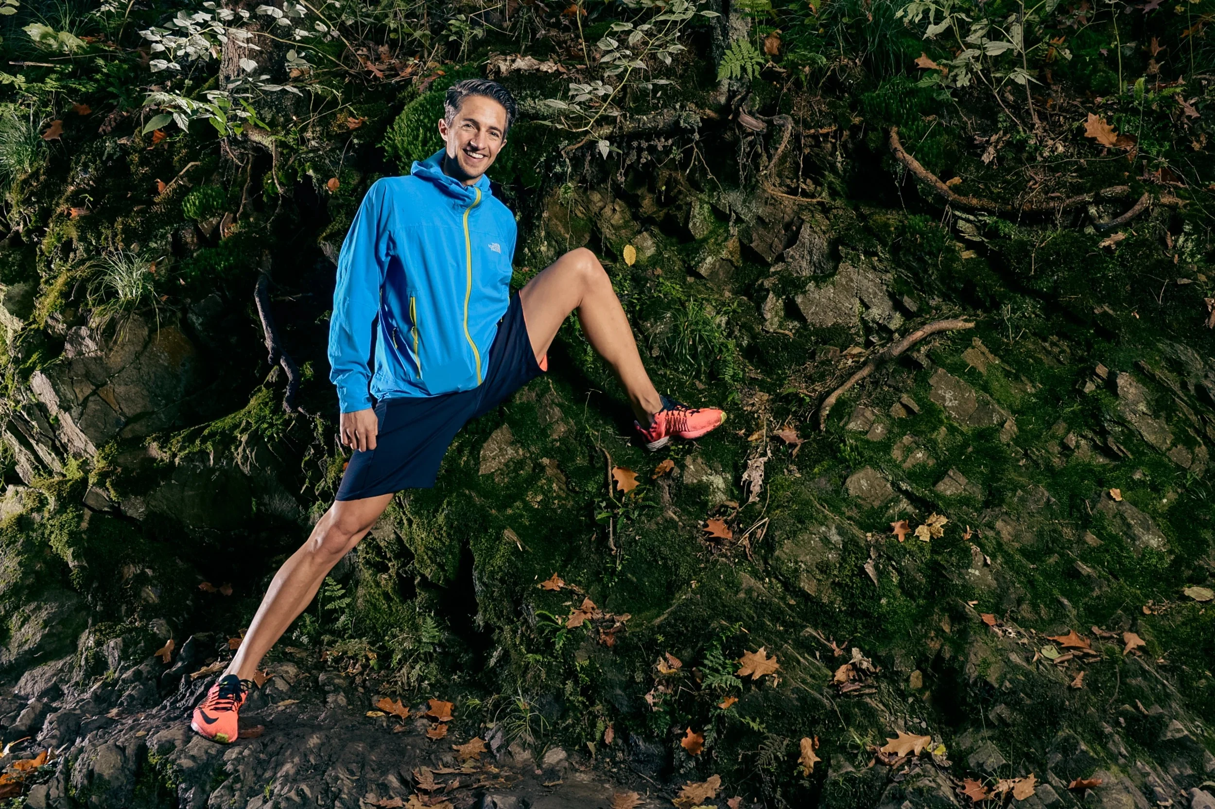 Male runner in a blue windbreaker stretching against a mossy forest wall.